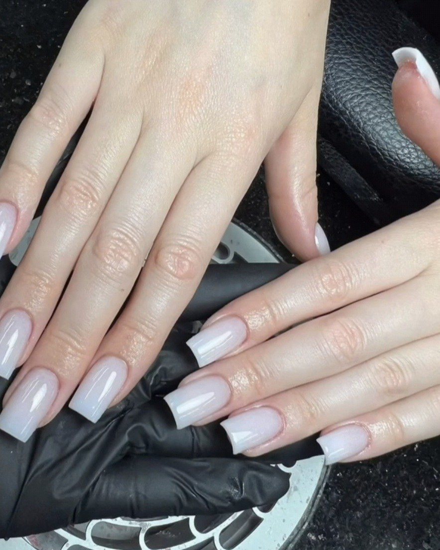 Lange, vierkante nagels in melkwit, de hand rust op een zwarte handschoen aan de nageltafel